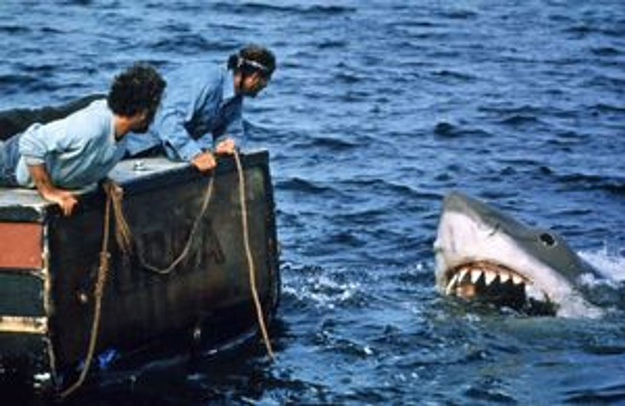 Richard Dreyfuss and Robert Shaw lean over the side of a boat as a large great white shark with open jaws emerges from the water in a scene from the film Jaws.