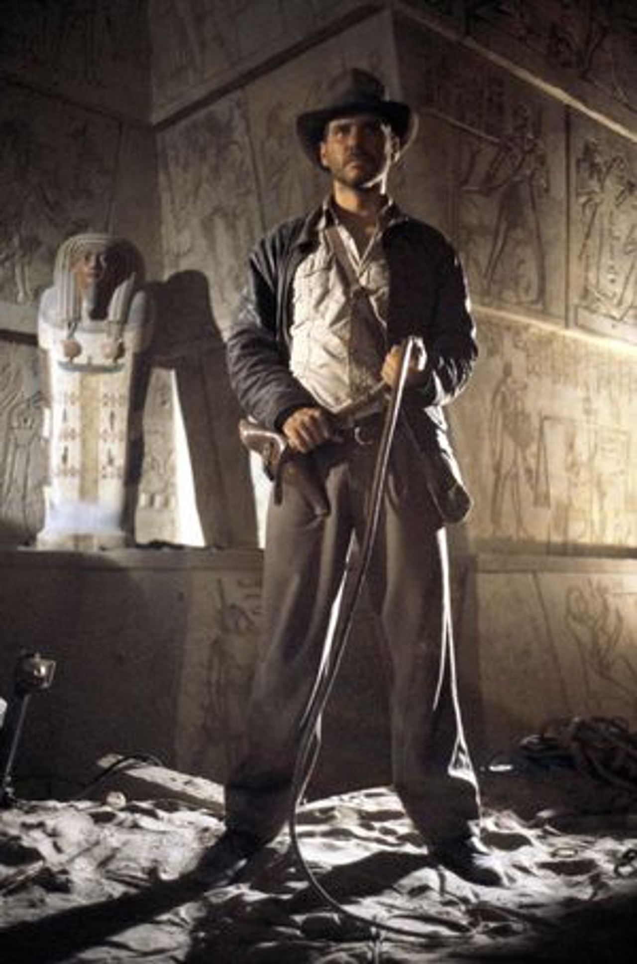 Harrison Ford as Indiana Jones stands holding a whip in an ancient, dimly lit chamber with Egyptian hieroglyphs and a sarcophagus in the background.