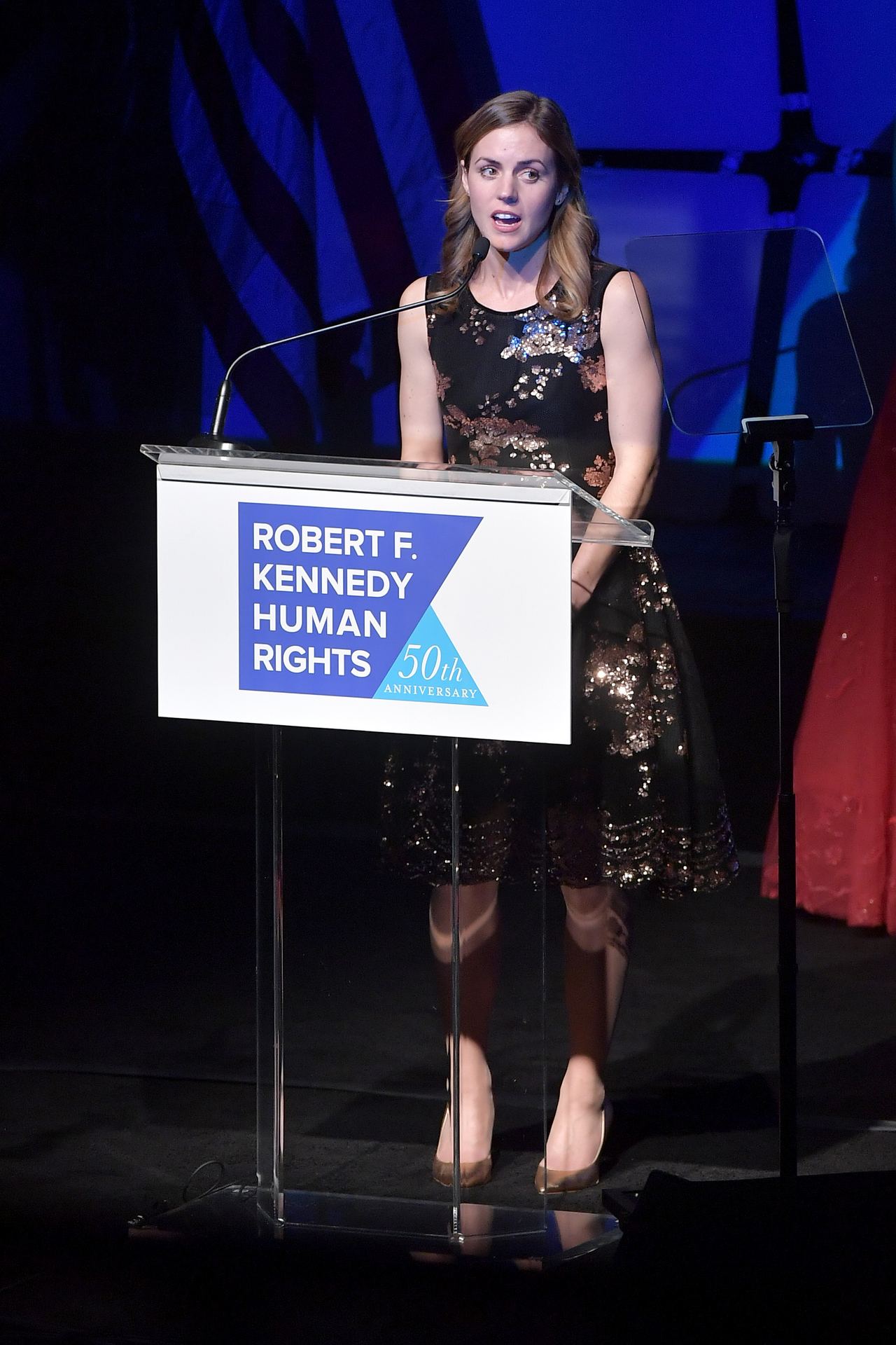 Kyle Kennedy Kerr addresses the audience from behind a podium at the 2019 Robert F. Kennedy Human Rights Ripple of Hope Awards on December 12, 2018, in New York City. Standing at the mic, she wears a sleeveless black knee-length dress adorned with shimmering gold and bronze sequins, paired with metallic pointed-toe heels. | Source: Getty Images