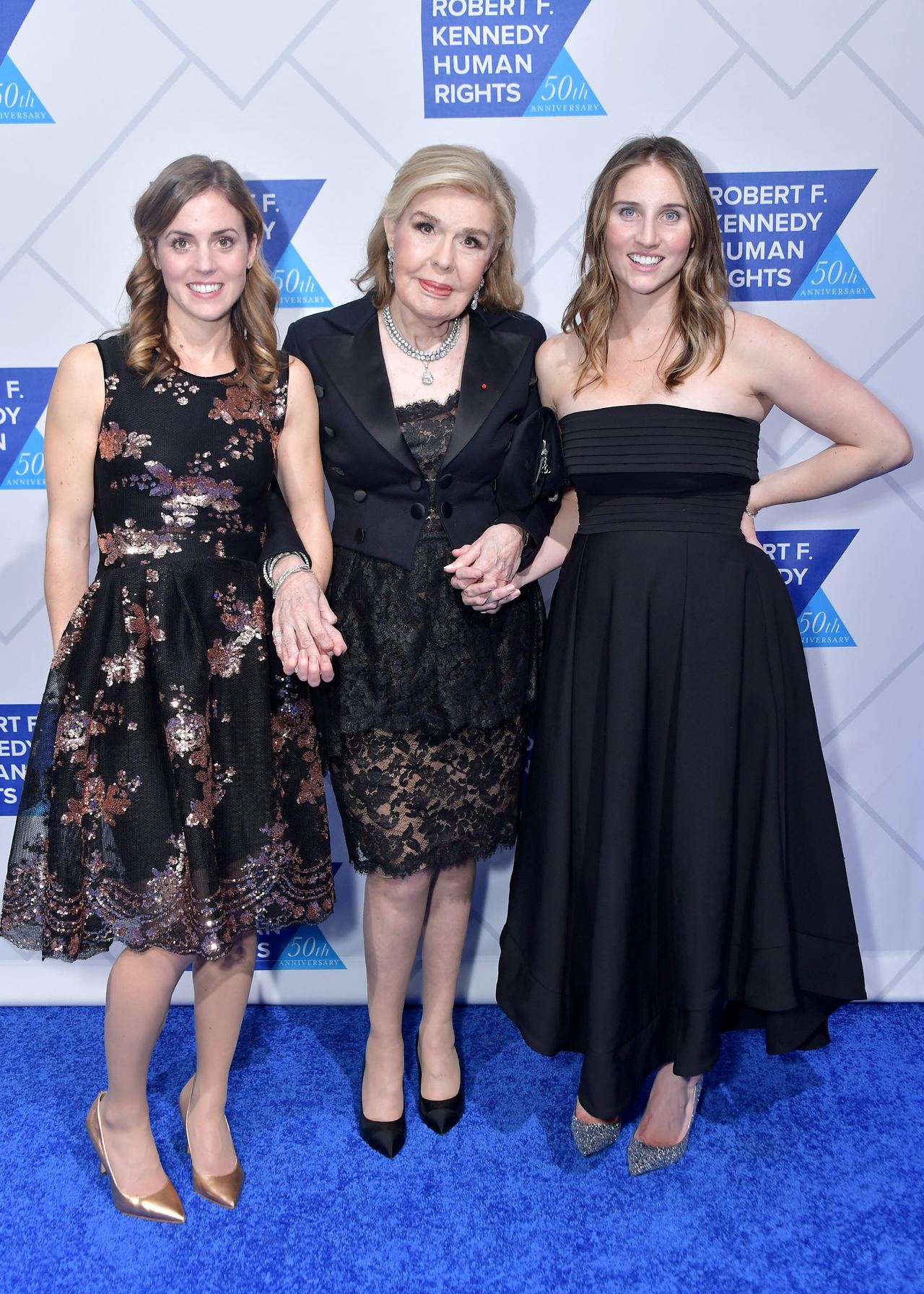 Kyle Kennedy Kerr, Marianna Vardinoyannis, and Rory Kennedy Camillo link hands on the blue carpet at the 2019 Robert F. Kennedy Human Rights Ripple of Hope Awards on December 12, 2018, in New York City. Kyle wears a sequined knee-length black dress with metallic pumps, standing alongside Marianna in an elegant black lace ensemble with a cropped jacket and pearl necklace, while Rory poses on the right in a strapless black pleated gown. | Source: Getty Images